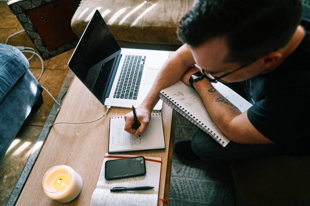 Man with a black shirt and glasses writing on a notepad with a laptop on the tab.e